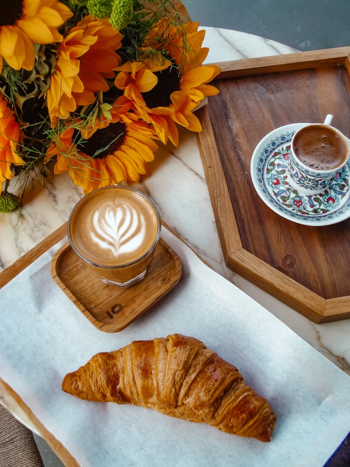 A cozy breakfast setup featuring a croissant, latte, espresso, and sunflower bouquet.